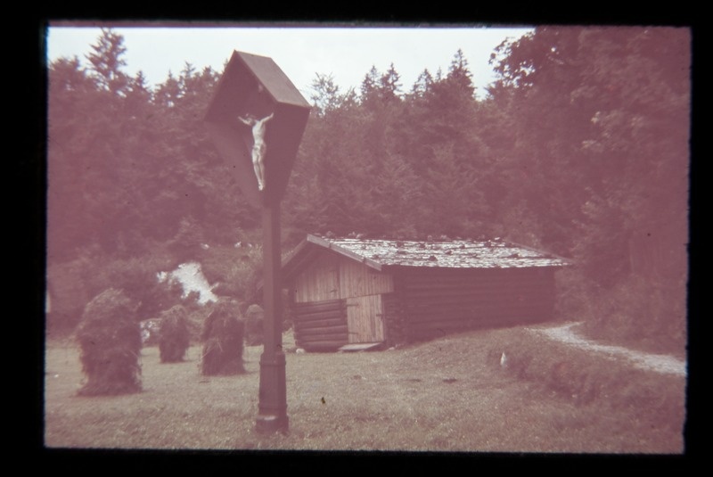 Katusega krutsifiks, eemal madal palkhoone, ümber puud.