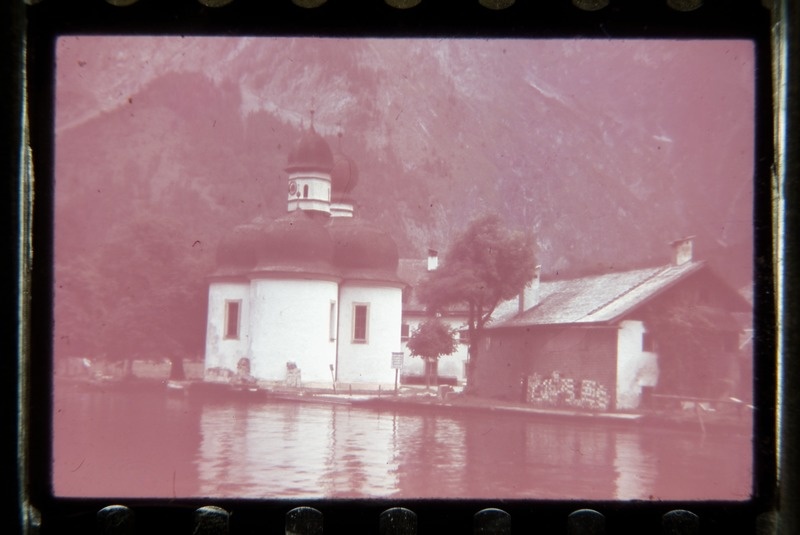 Kuppelkatustega väike kirik ja hooned veekogu ääres, taga mäenõlv.