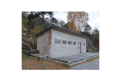 Näkymä Vallisaaren itärantaa kulkevalle tielle. Se on retkisataman tuntumassa jo suurin piirtein Kuninkaansaarta vastapäätä. Punatiilinen rakennus vasemmalla liittyy saaren torpedoluolaston toimintaan. Rakennus on rapattu harmaalla laastilla, josta osa on karissut pois. Rakennuksen päätyä vasten nojaa lavoja ja julkisivun edustalle on rakennettu alusta. Äärimäisenä oikealla pilkottaa meri. rephoto