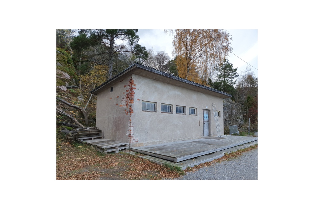 Näkymä Vallisaaren itärantaa kulkevalle tielle. Se on retkisataman tuntumassa jo suurin piirtein Kuninkaansaarta vastapäätä. Punatiilinen rakennus vasemmalla liittyy saaren torpedoluolaston toimintaan. Rakennus on rapattu harmaalla laastilla, josta osa on karissut pois. Rakennuksen päätyä vasten nojaa lavoja ja julkisivun edustalle on rakennettu alusta. Äärimäisenä oikealla pilkottaa meri. rephoto