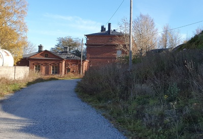 Näkymä Vallisaaresta. Oikealla on luotsitalo ja keskellä näkyy luotsitalon saunan ja pesutuvan rakennus. Vasemmalla alunperin venäläisten varuskuntansa saunaksi rakentama talo. Se on nykyään jo purettu. Tie rakennusten luo on säilynyt. rephoto