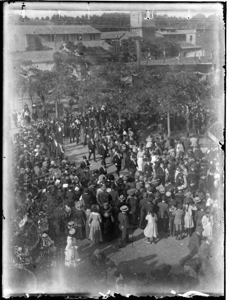 [First stone of the Post Office] - Act laying the foundation stone of the Post and Telegraph building. Arrival of the authorities on the site where the future building must be built on Avenida Ramon Folch. A band of music and the authorities are observed making their way throught the crowd. In the background is the Fire and Slaughterhouse building.
