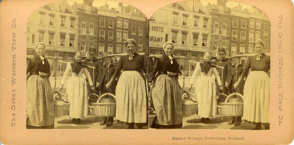 Een stereofoto van drie marktvrouwen die manden dragen met een juk. Op de achtergrond een aantal Nederlandse pakhuizen. Onder de foto: "Market Women, Rotterdam, Holland."