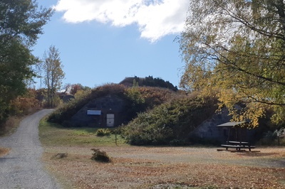 Näkymä Vallisaaren linnoituksilta saaren luoteisrannikolla. Maastoon on rakennettu kiviseinäisiä ruutivarastoja, jotka venäläiset rakensivat 1860-1870-luvuilla. Ne on katettu maakerroksella, ja ne saavat maaston kumpuilemaan. Hiekkainen kävelytie kiemurtelee kumpujen välistä. Vasemmalla näkyy holvikaarimainen betoninen sisäänkäynti varastoon. rephoto