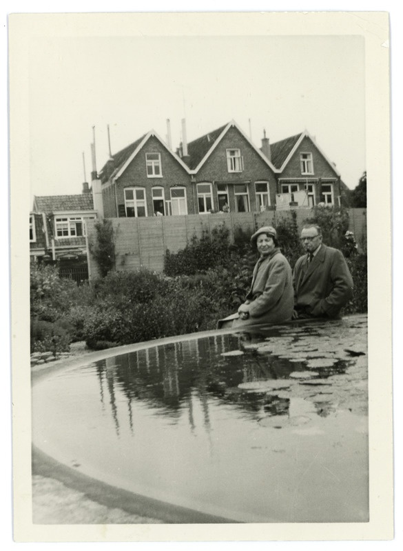 Mees ja naine Groningeni botaanikaaias veekogu äärel istumas, taustal elumajad