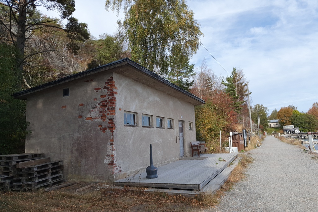 Näkymä Vallisaaren itärantaa kulkevalle tielle. Se on retkisataman tuntumassa jo suurin piirtein Kuninkaansaarta vastapäätä. Punatiilinen rakennus vasemmalla liittyy saaren torpedoluolaston toimintaan. Rakennus on rapattu harmaalla laastilla, josta osa on karissut pois. Rakennuksen päätyä vasten nojaa lavoja ja julkisivun edustalle on rakennettu alusta. Äärimäisenä oikealla pilkottaa meri. rephoto