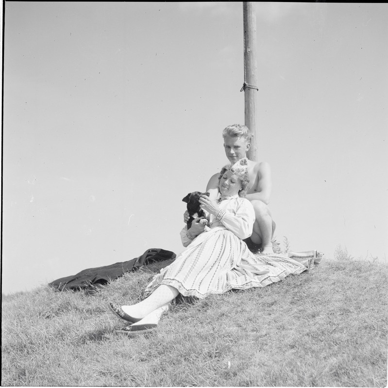 Naine Muhu rahvariietes - proua Naaber, süles koer, mees seljataga.