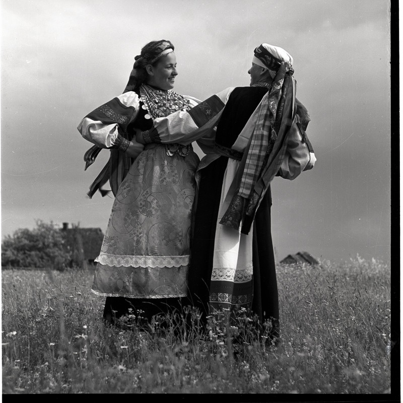 Setu rahvariietes Olli Kalju ja Anni Kalju tantsivad, tagaplaanil taluhooned