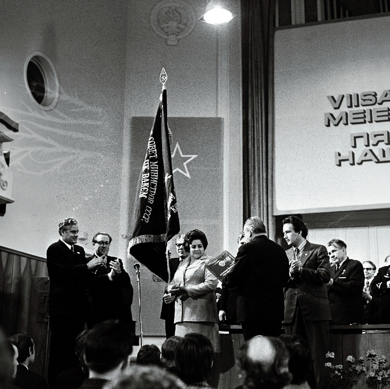 1974. a. üleliidulise sotsialistliku võistluse tulemuste põhjal Tallinnale määratud NLKP Keskkomitee, NSV Liidu Ministrite Nõukogu, ÜAÜKN-i ja ÜLKNÜ Keskkomitee rändpunalippu hoiab sotsialistliku töö kangelane, Tallinna linna aukodanik A. Valdov. Vasakult 1. EKP Tallinna Linnakomitee esimene sekretär N. Johanson, 2. Tallinna TSN Täitevkomitee esimees J. Kallion, 6. EKP Keskkomitee esimene sekretär J. Käbin.